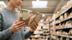 Person vergleicht zwei Lebensmittelpackungen in einem hellen Supermarktgang
