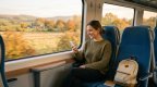 Lächelnde junge Frau sitzt am Fenster eines hellen Fernzugs und schaut auf ihr Smartphone, neben ihr ein Rucksack, draußen eine sonnige Herbstlandschaft