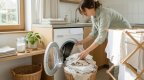Hände legen sortierte helle Wäsche in eine geöffnete Waschmaschine in einem hellen Waschraum, daneben ein Wäscheständer mit trocknenden Handtüchern im Sonnenlicht
