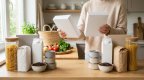 Hände halten zwei schlichte Müslipackungen zum Vergleich über einer Küchenarbeitsplatte mit unbeschrifteten Nudelgläsern, Milchkartons, Dosen und Kaffeebohnen