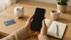 Hände halten ein Smartphone mit dunklem Bildschirm über einem hellen Schreibtisch mit unbeschrifteter Bankkarte, Sparschwein, Notizbuch und Kaffeetasse im Morgenlicht