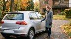 Frau im grauen Wollmantel steht mit Autoschlüssel in der Hand neben einem silbernen Kompaktwagen in einer herbstlichen Einfahrt mit Laub im Morgenlicht