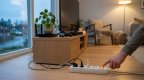Hand schaltet eine weiße Steckdosenleiste neben einem TV-Sideboard mit Router, Fernseher und Spielcontroller in einem gemütlichen Wohnzimmer am Abend aus