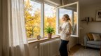 Person im Strickpullover mit Teetasse steht am weit geöffneten Fenster eines hellen Wohnzimmers, darunter ein Heizkörper, auf der Fensterbank Hygrometer und Grünpflanze, draußen Herbstbäume
